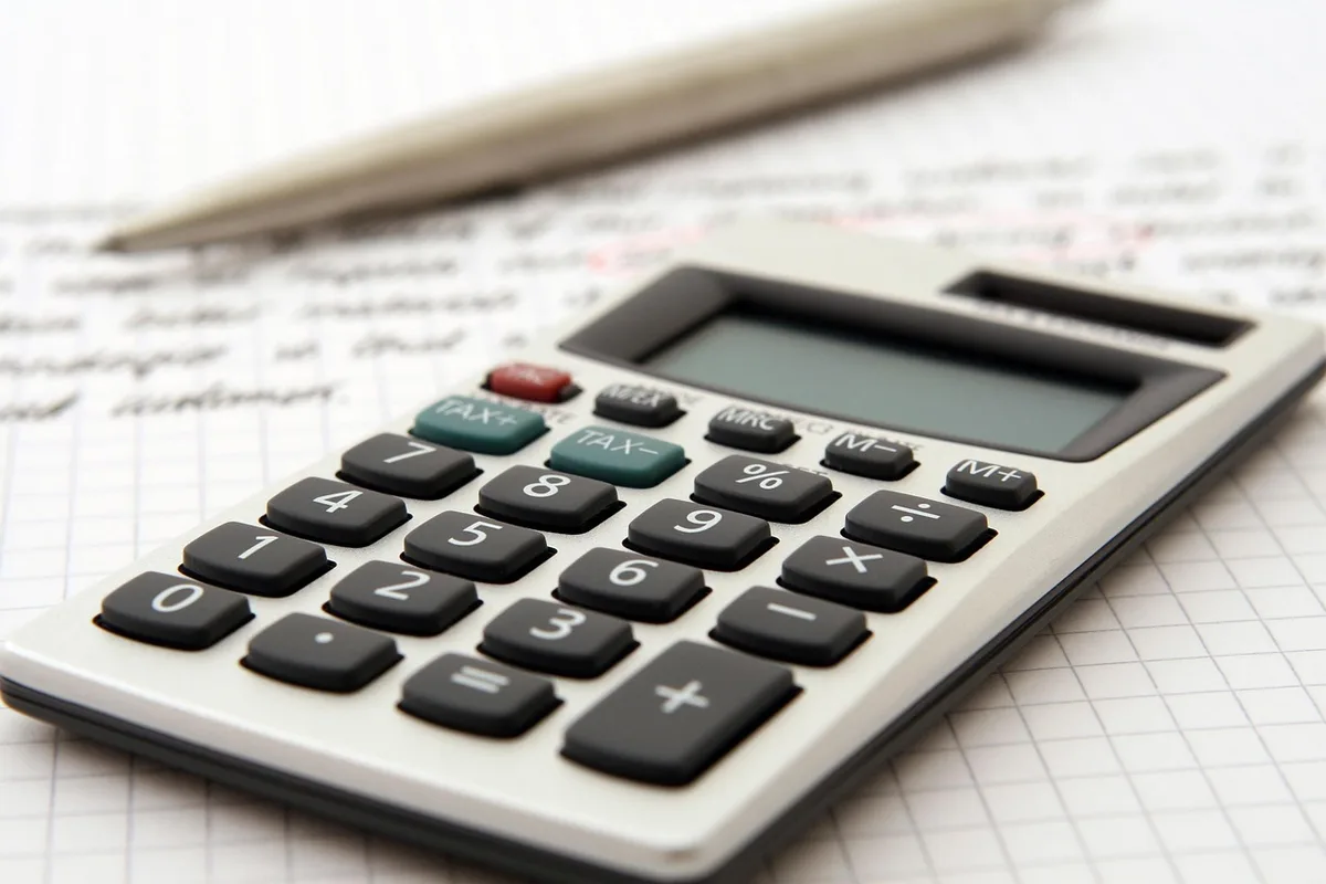 Main écrivant un budget à côté d'une calculatrice et de factures sur une table ensoleillée